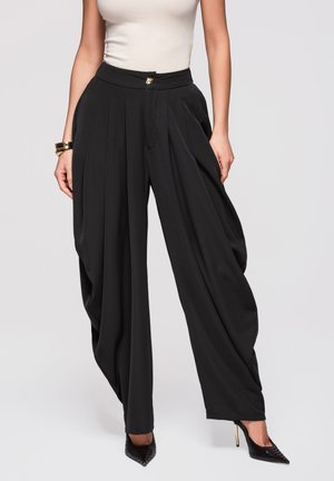 Femme portant un pantalon noir à taille haute, large et plissé, des talons noirs à bouts pointus, un haut blanc sans manches et un bracelet doré au poignet gauche.