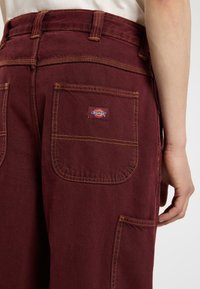 Pantalones cargo burdeos con costuras amarillas en contraste, bolsillos traseros visibles y un pequeño parche de logo en el bolsillo derecho.