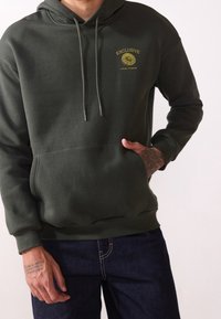 Hombre con sudadera con capucha verde oscuro con texto amarillo "EXCLUSIVE LOCAL FLAVOR" y gráfico de limón, con una mano en el bolsillo, combinado con jeans oscuros.