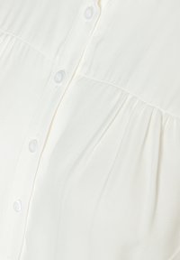 White shirt featuring a button-down design, textured fabric, and subtle pleating detail below the buttons. Smooth surface and lightweight.