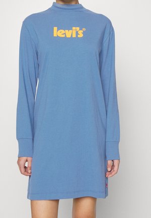 Person iført en blå minikjole med lange ærmer og gul "levi's"-logo på brystet mod en ensartet lys baggrund.