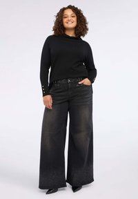 Maglione nero a maglia con collo a cascata e dettagli con bottoni sulle maniche, abbinato a jeans neri a gamba larga con un sottile effetto ombre.