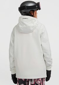 Witte, waterdichte skijas met een capuchon. Voorzien van elastische manchetten en een logo op de rug. Draagt samen met gestreepte sneeuwbroeken en zwarte handschoenen.