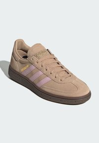 Beige suède sneaker met drie roze strepen, een bruine rubberen zool en gouden branding op de tong. Klassiek low-top ontwerp met veters.