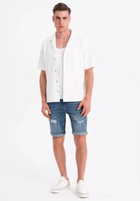 Vit kortärmad skjorta med knappar, bärs över en vit linne, tillsammans med blå slitna denimshorts och vita sneakers.