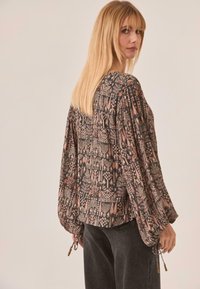 Blusa negra y estampada con mangas largas y amplias, puños elásticos y lazos de cuentas. Presenta diseños florales y geométricos en tonos apagados.