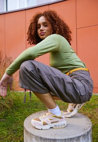 Långärmad ljusgrön topp med präglad logotyp, grå cargobyxor och krämfärgade sneakers med rosa och gula detaljer, poserande på en betongcylinder.