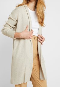 Beige ribbad kardigan med öppen front, långa ärmar och sidsprund, bärs över en vit skjorta och ljusbruna byxor.