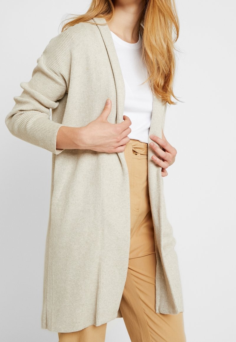 Beige ribbad kardigan med öppen front, långa ärmar och sidsprund, bärs över en vit skjorta och ljusbruna byxor.