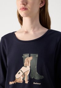 Navyblaues Langarm-T-Shirt mit einem Grafikmotiv eines Hundes und grünen Gummistiefeln. Das Geschirr des Hundes ist cremefarben mit einem runden Akzent.
