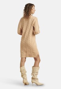 Abito maglione in morbido beige con motivi a trecce, maniche lunghe, spacchi laterali e orlo a costine. Abbinato a stivali beige al ginocchio.