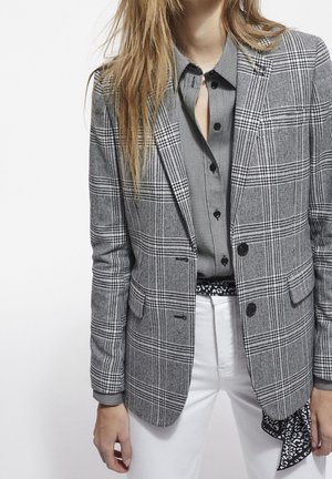 Femme portant un blazer et une chemise à carreaux noir et blanc avec un pantalon blanc, debout devant un fond blanc uni.