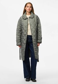 Chaqueta larga acolchada en verde apagado, con un amplio collar, cierres a presión y una cintura con correa. Combinada con jeans oscuros y zapatos negros.