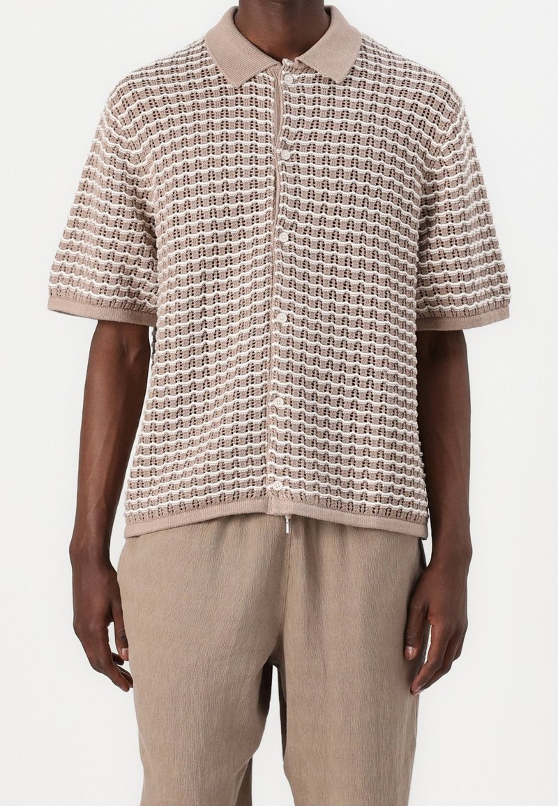 Homme portant un polo en maille beige à manches courtes avec boutons sur le devant et motif carré texturé, associé à un pantalon beige ample.