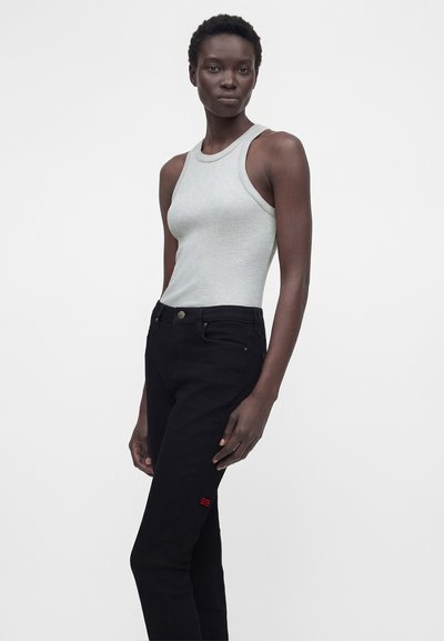 Γυναίκα που φοράει ανοιχτό γκρι αμάνικο ριμπ τοπ και ψηλόμεσα μαύρα τζιν με μικρό κόκκινο λογότυπο στον μηρό, στέκεται μπροστά σε λευκό φόντο.