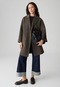 Vrouw in een donkerbruine oversized wollen jas, met wijde donker gekleurde spijkerbroek met omslag, loafers en een zwarte trekkoord handtas, tegen een effen achtergrond.