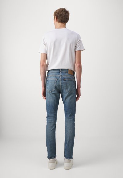 Jean bleus ajustés en denim, avec un design classique à cinq poches et une étiquette en cuir beige à l'arrière de la taille. T-shirt blanc porté.