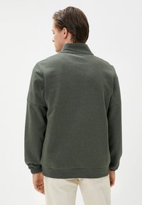 Sudadera verde oliva con cuello alto, puños elásticos y dobladillo. Textura de tela suave, corte relajado y diseño minimalista.