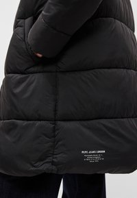 Črna napihnjena jakna ima kvadratne šive, gladko teksturo in stranske žepke. Bele besede za blagovno znamko so prikazane na spodnji levi strani.