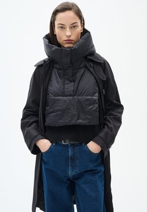 Černá péřová bunda s vysokým límcem, přes černý svetr, v kombinaci s tmavě modrými džíny. Minimalistický design.