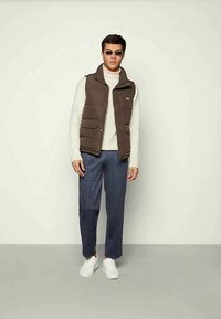 Brown puffer vest over a beige turtleneck, paired with blue patterned wide-leg trousers and white sneakers. Accent sunglasses complete the look.