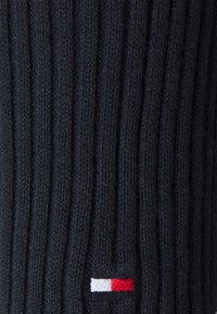 Dunkelblaue gerippte Strickware mit einem kleinen rechteckigen Logopatch, der weiße und rote horizontale Blöcke nahe der unteren Mitte zeigt.