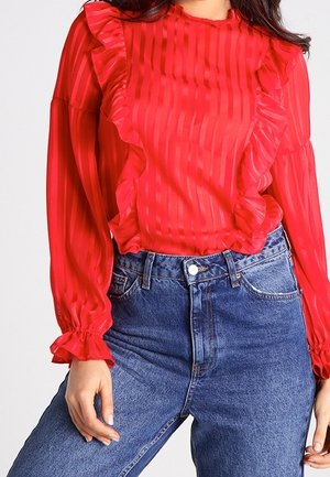 Kvinde iført en rød, gennemsigtig, stribet bluse med flæser foran og ved ærmekanterne, kombineret med højtsiddende blå jeans.