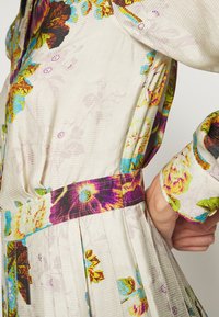 Květinové šaty z krémové látky s barevnými akcenty. Mají plisovaný design a odpovídající pásek v pase pro strukturu.