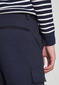 Personne portant un pantalon bleu marine avec une poche arrière et une poche cargo, associé à un haut à manches longues rayé bleu marine et blanc.