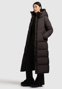 Pikk must puffertakki kõrge kraega, millel on tikitud paneelid, lukuga ees ja küljetaskud, kantud musta pluusi ja pükste peal.