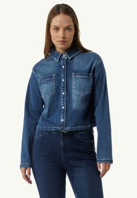 Kurz geschnittene Jeansjacke in Dunkelblau mit spitzem Kragen, Knopfleiste, zwei Brusttaschen und einer Bindeband-Detail am Saum.