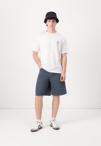 Camiseta blanca con un pequeño gráfico rosa, pantalones cortos azules, sombrero tipo cubo negro y zapatillas blancas con rayas negras. Conjunto informal sencillo.