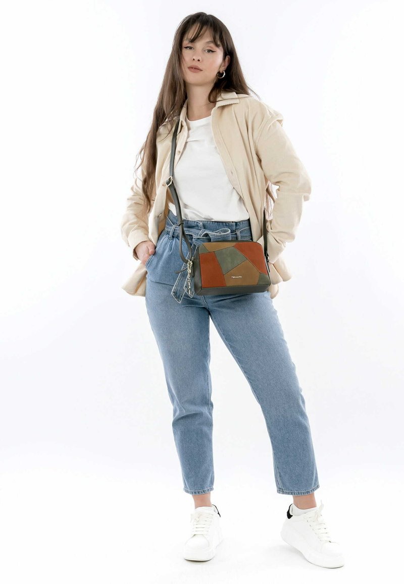 Meer-kleurige schoudertas gemaakt van leer met geometrisch patchwork; voorzien van verstelbare riem en ritssluiting. Model draagt een beige jas en spijkerbroek.
