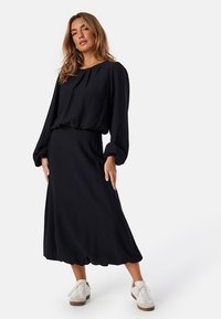 Svart långärmad klänning med lös passform, samlad halsringning och en lätt utsvängd kjol, tillsammans med vita sneakers och ett texturerat tyg.