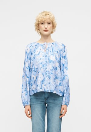 Žena so krátkymi kučeravými blond vlasmi, oblečená v svetlomodrej blúzke s kvetinovým vzorom a modrých džínsoch, stojaca na bielom pozadí.