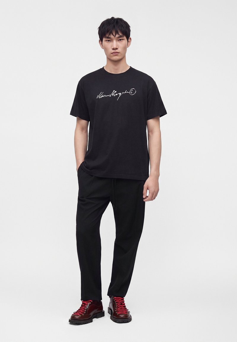 Jeune homme debout portant un t-shirt graphique noir, un pantalon noir et des bottes rouge foncé avec des lacets rouge vif sur un fond blanc uni.