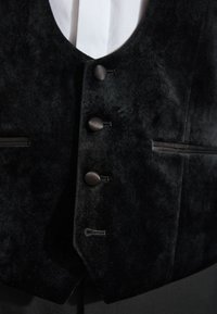 Gilet en velours noir avec quatre boutons recouverts de tissu assortis et poches passepoilées sur une chemise blanche.