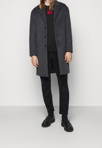 Cappotto grigio in lana con rever a punte, tre bottoni frontali, abbinato a un maglione nero, pantaloni di velluto a coste neri e mocassini neri.