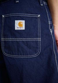 Jeans de color azul marino con un bolsillo trasero que tiene costuras blancas y un parche cuadrado blanco con el logotipo de Carhartt en naranja.