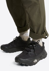 Chaussures de sport noires avec semelles en caoutchouc texturées, tige en mesh et matière synthétique. Associées à un pantalon vert olive et des chaussettes crème portant un logo.