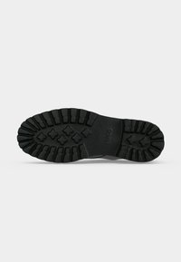 Semelle de chaussure en caoutchouc noir avec des crampons profonds et des motifs en forme de croix, centre texturé, et logo de la marque embossé au centre.