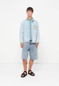 Light blue denim jacket with a chest pocket, paired with matching denim shorts. Black sandals with straps, and a teal t-shirt underneath.