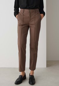 Bruine slim fit broek met zijzakken, voorzijde met vouwdetails en een gladde textuur. Gecombineerd met zwarte loafers.