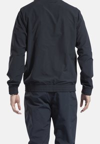 Man med en svart jacka med elastiska muddarmar och matchande byxor, som står med avslappnade armar, vänd bort från kameran mot en enfärgad bakgrund.