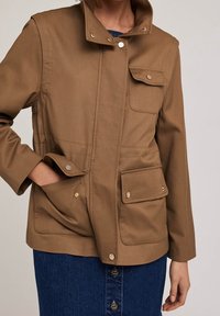 Brown cotton jacket with a stand collar, snap buttons, and four pockets. Features a straight cut and a textured fabric.