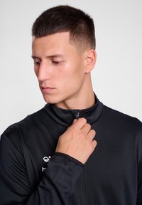 Zwarte sportjack met rits en hoge kraag, ontworpen voor een slim fit en gemaakt van textuurstof. Voorzien van een logo op de linkerborst.