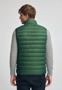 Grüne Pufferweste mit vertikalen Steppabschnitten. Hoher Kragen und leichtes Material. Über einem strukturierten grauen Pullover getragen.