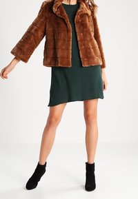 Brun fuskpälsjacka med en fyrkantig form, texturerat randigt mönster och vida ärmar, tillsammans med en grön klänning och svarta ankelstövlar.