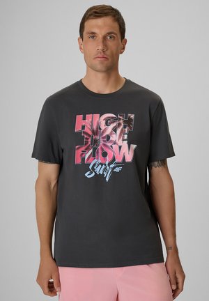 Mężczyzna w czarnej koszulce z różowo-niebieskim napisem "High Tide Flow Surf" i różowych szortach na szarym tle.