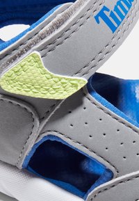 Grå sneakers med blå detaljer, med en texturerad gulgrön klaff, perforerade sidor och ett kardborreband. Mjuk inredning synlig.